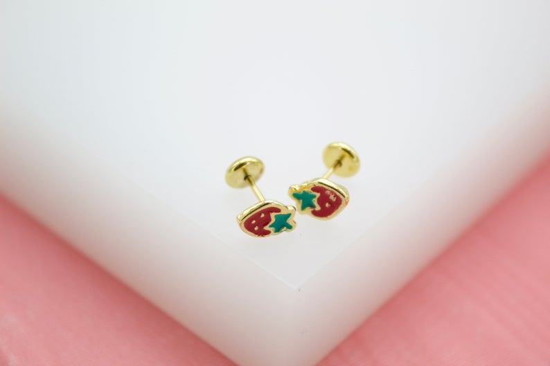 A pair of gold stud earrings featuring playful red and green fish designs rests on a white surface.