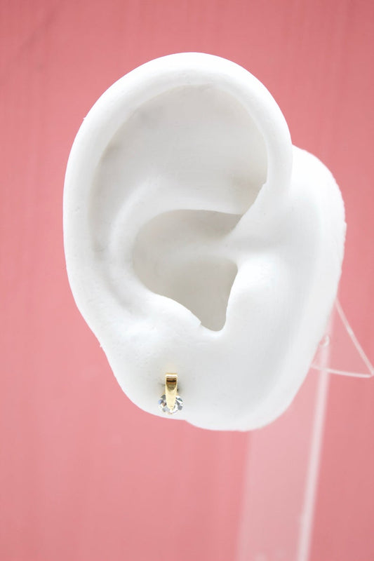 A white mannequin ear displays a small gold stud earring featuring a sparkling cubic zirconia stone against a pink background.