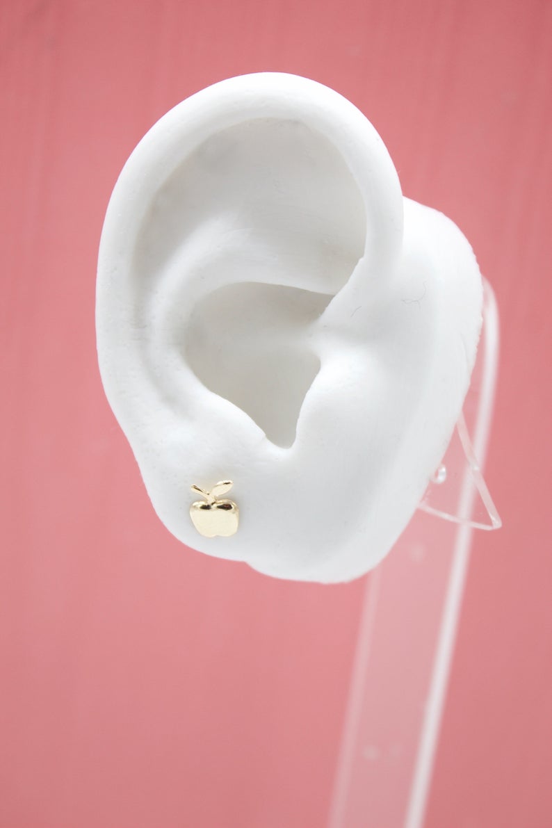 A white mannequin ear displays a small, shiny gold apple stud earring against a pink background.
