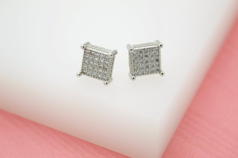 A pair of silver square stud earrings encrusted with sparkling cubic zirconia stones rests on a white display block.