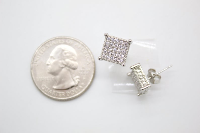 A pair of pave-set square diamond stud earrings rests next to a US quarter coin for scale on a light surface.