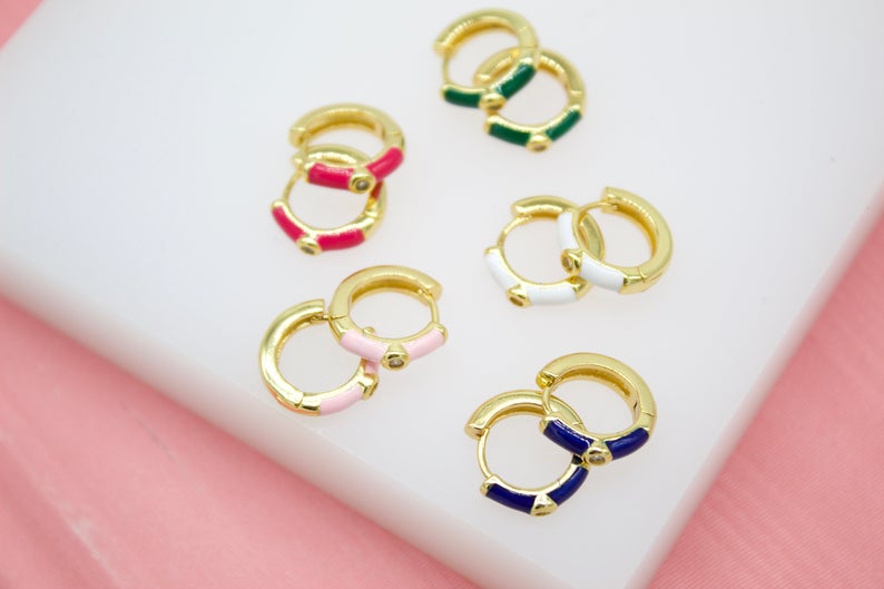 Several pairs of colorful enamel gold huggie hoop earrings with sparkling rhinestones rest on a white display surface.