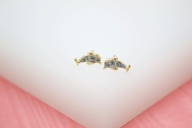 A pair of gold dolphin stud earrings features sparkling cubic zirconia pave details against a white and pink background.