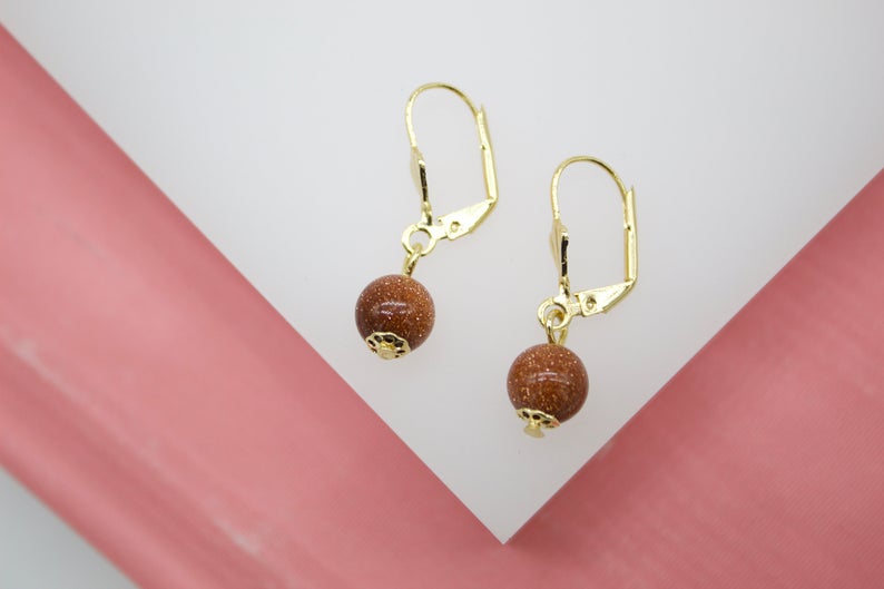 A pair of gold leverback earrings features dangling brown sandstone bead charms against a pink and white background.