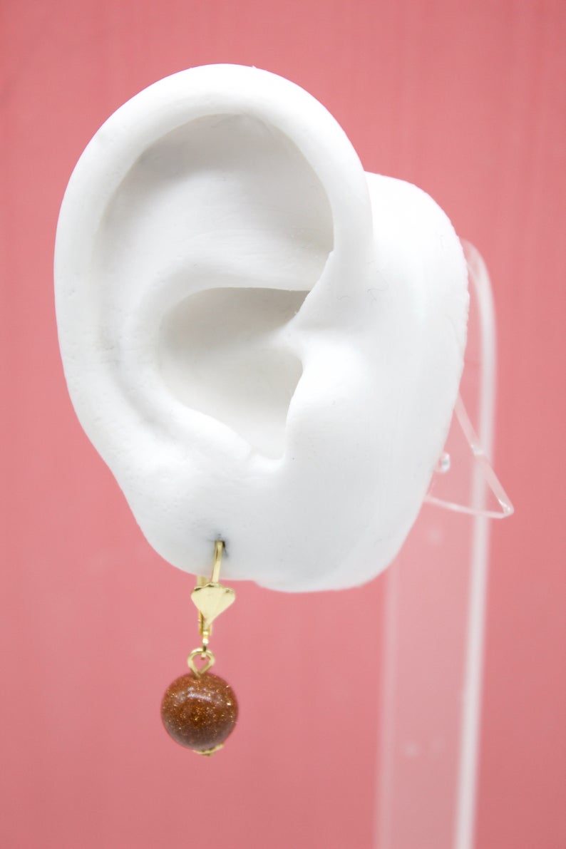 A white model ear displays a gold leverback earring featuring a dangling brown sandstone gemstone bead against a pink backdrop.