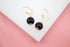 A pair of gold leverback earrings features glossy black sphere beads resting on a white display surface against a pink background.