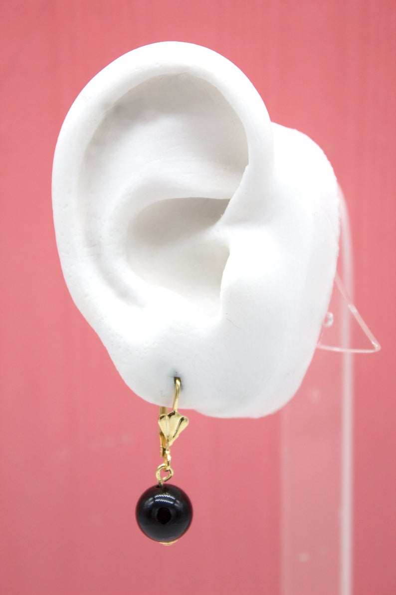 A white mannequin ear displays a gold leverback earring featuring a dangling black bead.