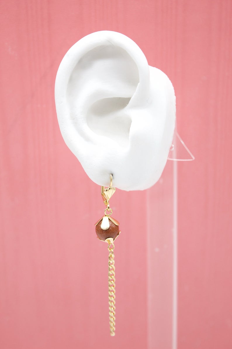 A white mannequin ear displays a gold dangle earring featuring a sparkling brown sandstone bead and a delicate chain drop against a pink background.