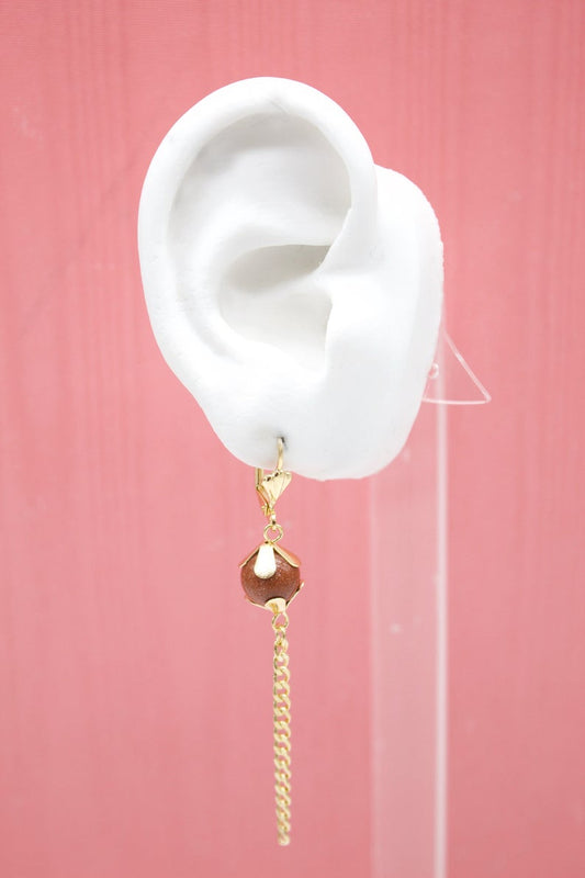 A white mannequin ear displays a gold dangle earring featuring a sparkling brown sandstone bead and a delicate chain drop against a pink background.