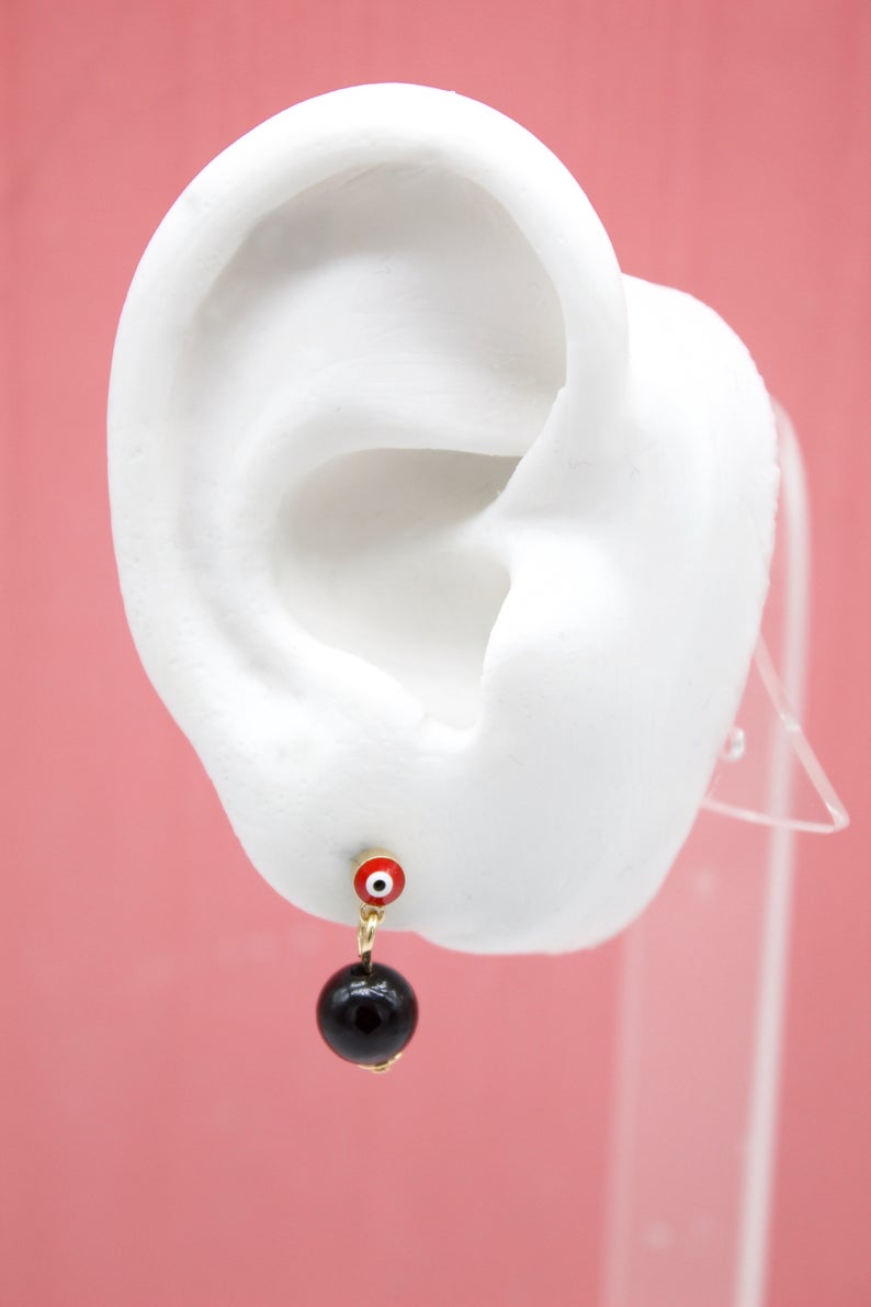 A white mannequin ear displays a gold evil eye stud earring with a hanging black bead against a pink background.