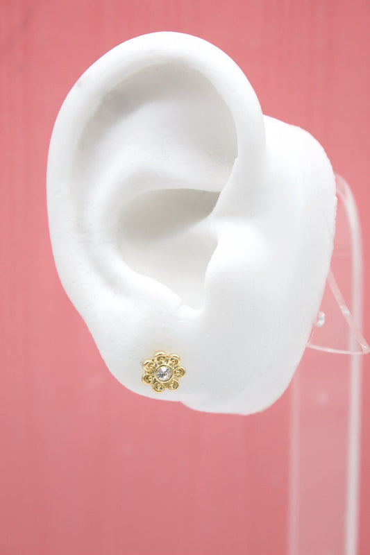 A white mannequin ear displays a dainty gold flower stud earring featuring a sparkling central rhinestone against a pink background.