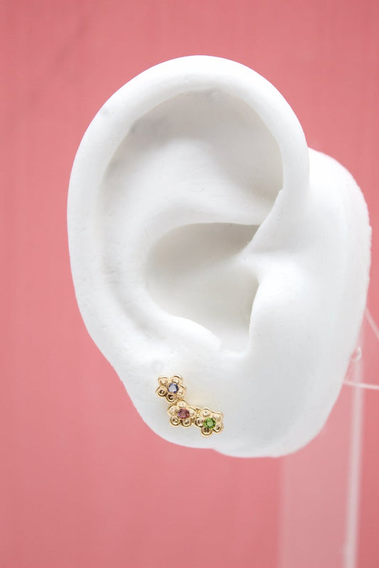 A white mannequin ear displays a delicate gold floral climber earring featuring colorful gemstone accents against a soft pink background.