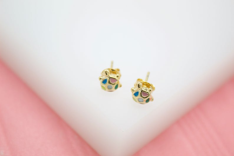 A pair of gold turtle stud earrings featuring colorful enamel details sits on a white display surface against a soft pink background.