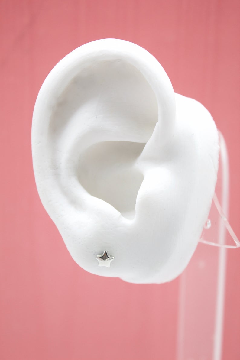 A white mannequin ear displays a tiny sterling silver star stud earring against a pink background.