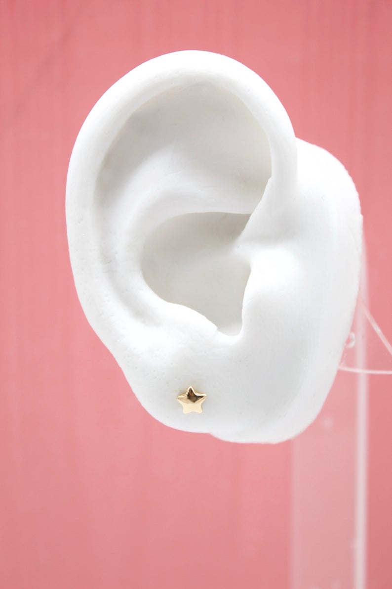 A white mannequin ear displays a tiny gold star stud earring against a pink background.