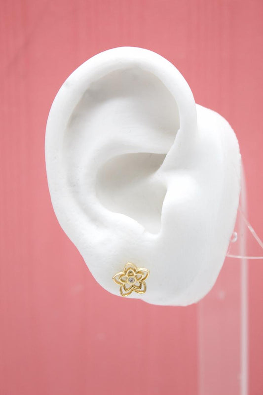 A white mannequin ear displays a gold flower stud earring featuring a central gemstone against a pink backdrop.