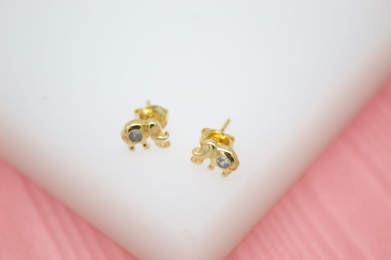 A pair of gold elephant stud earrings featuring sparkling cubic zirconia stones rests on a white display surface against a soft pink background.