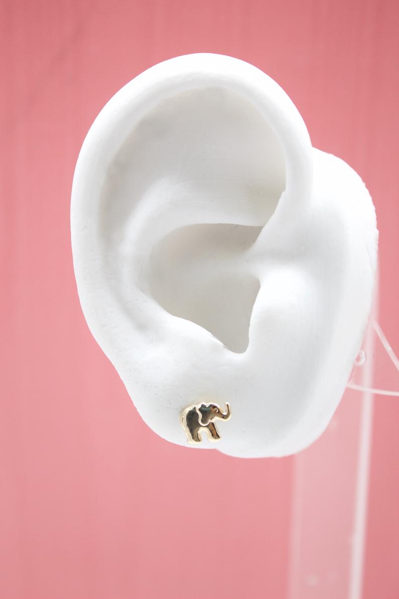 A white mannequin ear displays a small gold elephant stud earring against a pink background.