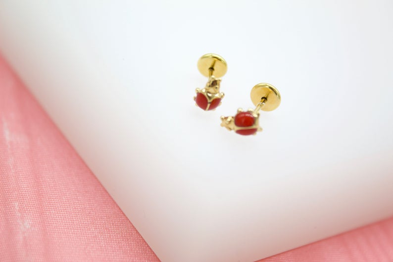 A pair of gold ladybug earrings with red enamel details rests on a white display surface.