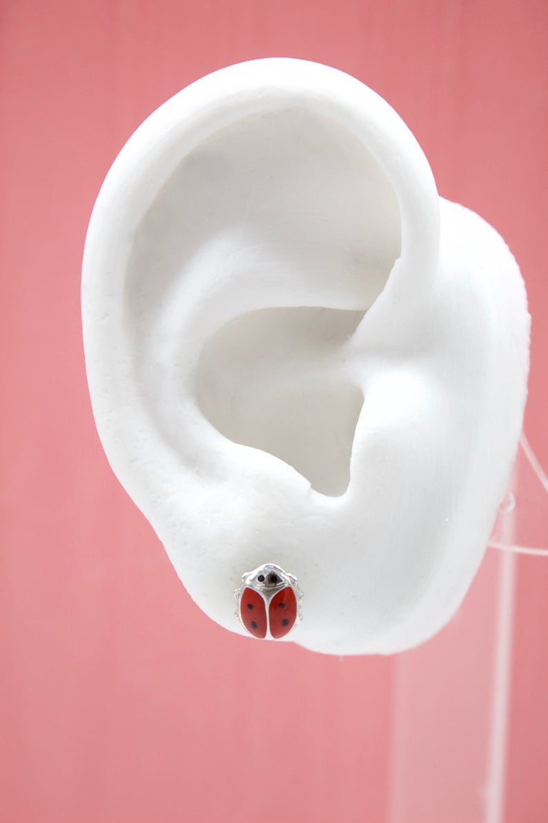 A white mannequin ear displays a cute red and black ladybug stud earring against a pink background.