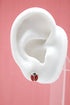 A white mannequin ear displays a cute red and black ladybug stud earring against a pink background.