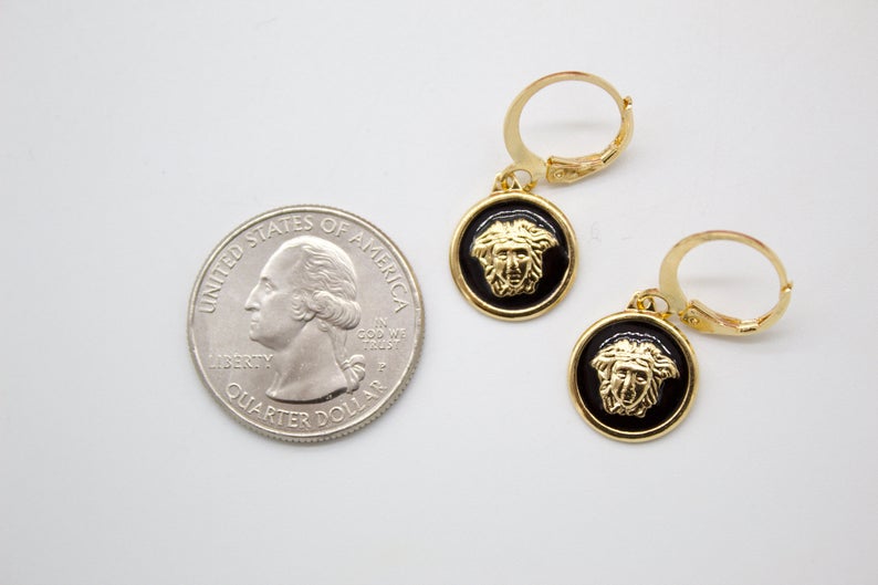 A pair of gold-plated leverback earrings featuring round black enamel pendants with the Medusa head emblem rests beside a US quarter for size comparison.