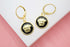 A pair of gold-plated leverback earrings features round pendants with a black enamel background and a raised Medusa head design.