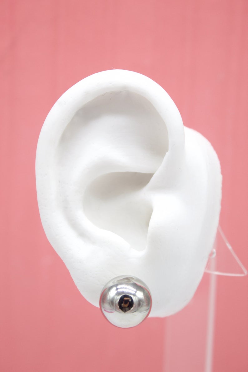 A white model ear displays a shiny silver ball stud earring against a pink background.