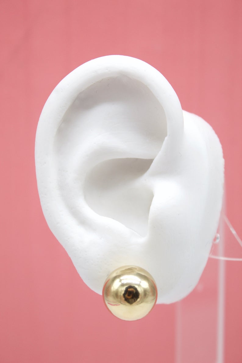 A white mannequin ear displays a shiny, spherical gold ball stud earring against a pink background.