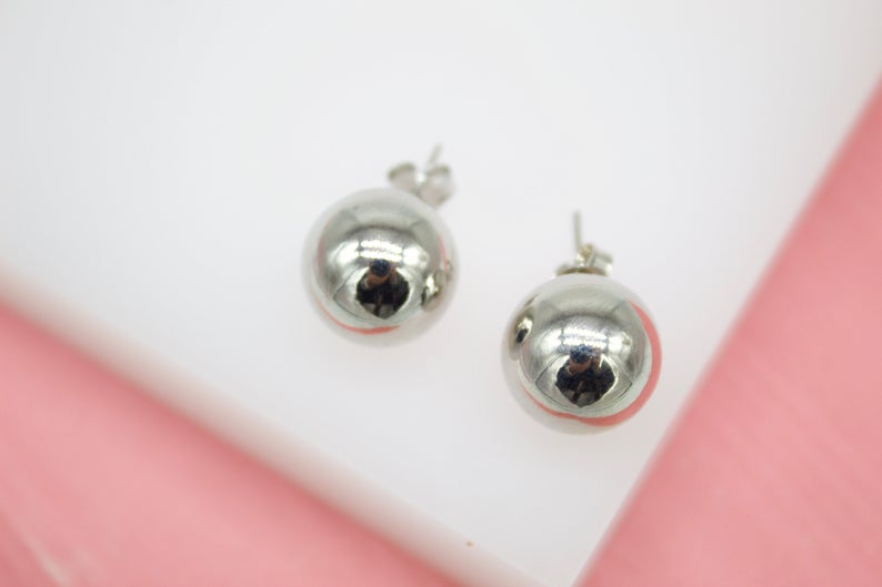 A pair of polished silver ball stud earrings rests against a white and pink backdrop.