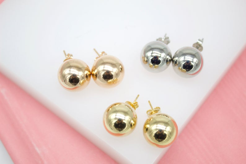Three pairs of glossy ball stud earrings in gold and silver finishes rest on a white display surface against a pink background.