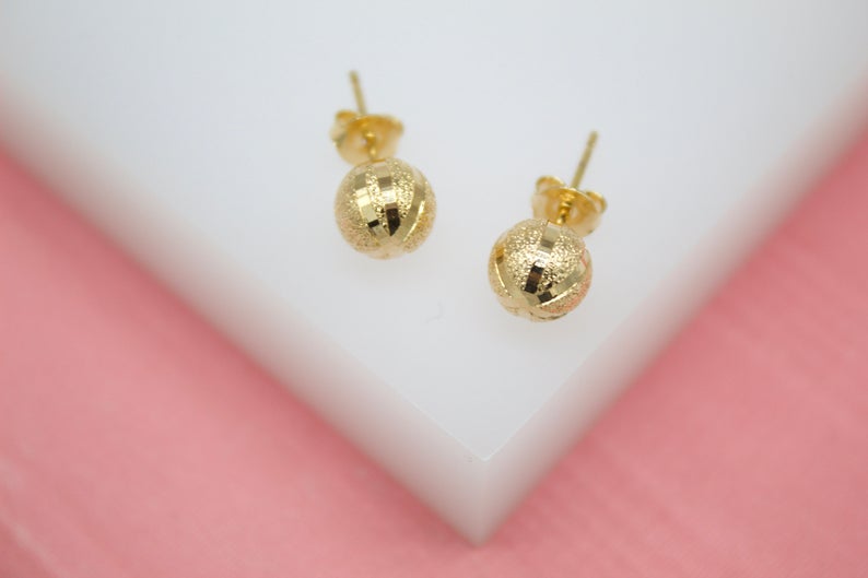 A pair of gold ball stud earrings featuring a textured, diamond-cut finish rest on a white surface against a soft pink backdrop.