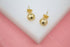 A pair of gold ball stud earrings featuring a textured, diamond-cut finish rest on a white surface against a soft pink backdrop.