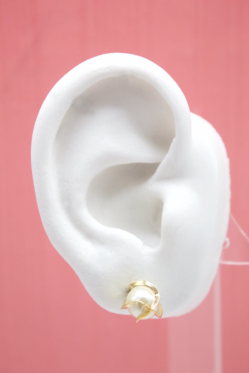 A white mannequin ear displays a gold and pearl tulip flower stud earring against a pink background.