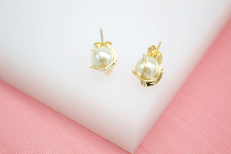 A pair of elegant gold stud earrings featuring lustrous faux pearls cradled in delicate metallic settings rests on a white display surface against a pink background.