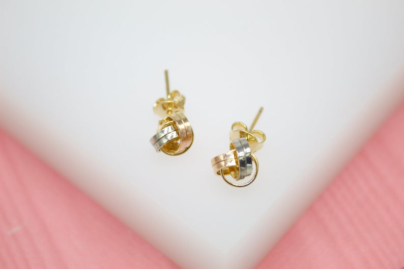 A pair of intertwined tri-color gold knot stud earrings rests on a white surface.