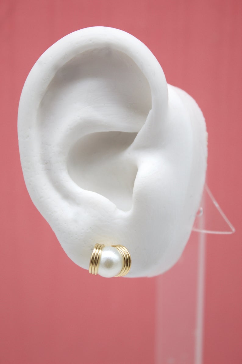 A white pearl stud earring with ribbed gold detailing sits in the lobe of a white mannequin ear against a solid pink background.