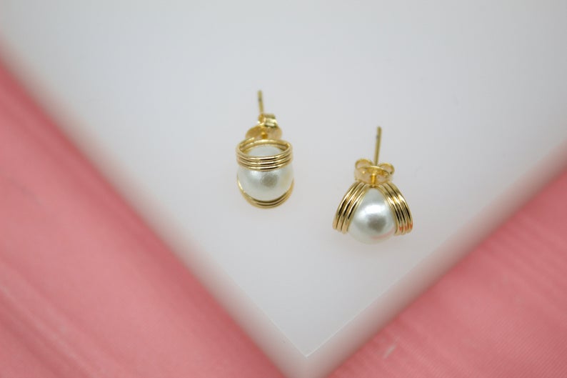 A pair of gold wire-wrapped pearl stud earrings rests on a white display stand against a soft pink background.