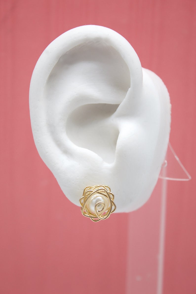 A white mannequin ear displays a delicate gold wire-wrapped pearl stud earring against a pink background.