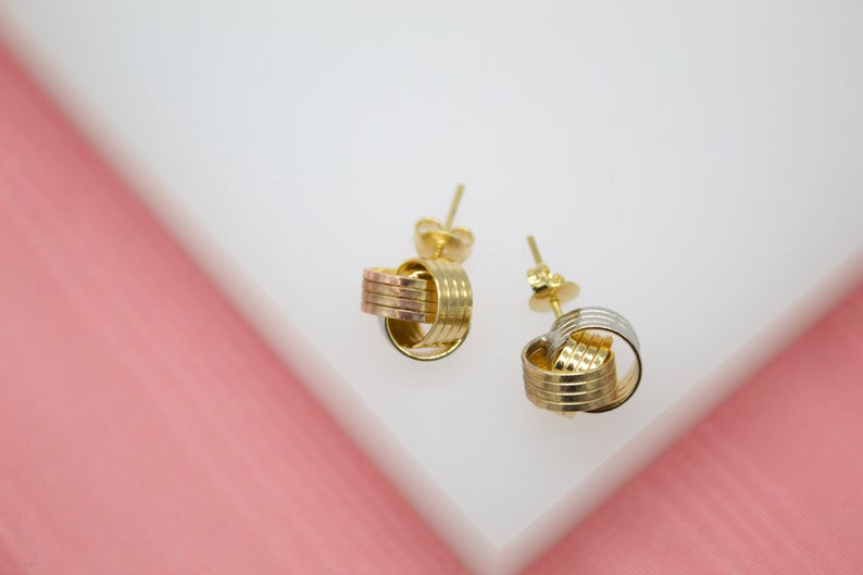 A pair of intricately intertwined tri-color gold hoop stud earrings rest on a white display surface.