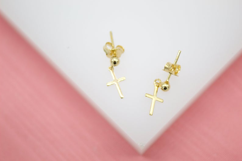 A pair of gold cross drop stud earrings rests on a white display against a pink background.