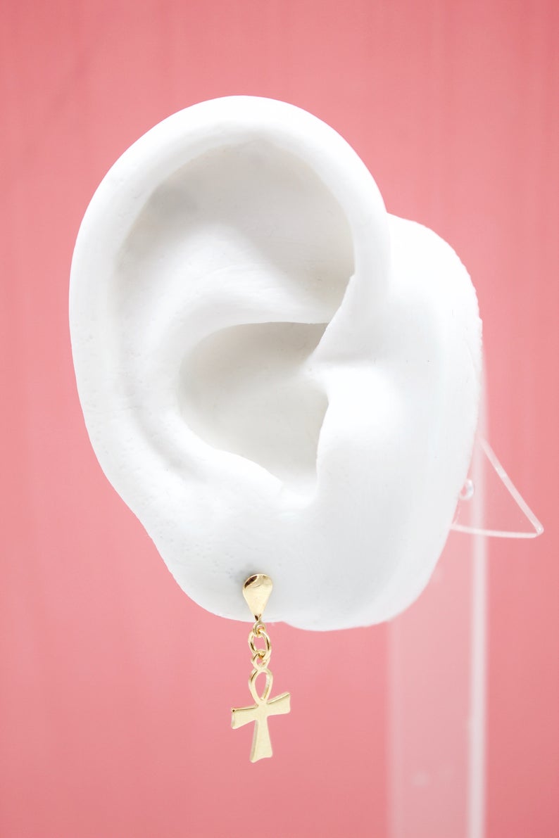 A white mannequin ear displays a delicate gold ankh cross dangle earring against a solid pink backdrop.