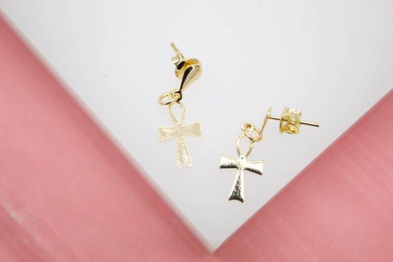 A pair of gold ankh drop earrings rests on a white display surface against a pink background.