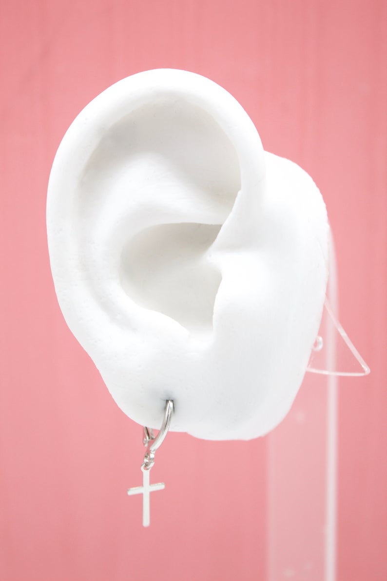 A white ear model displays a silver cross charm huggie hoop earring against a pink background.