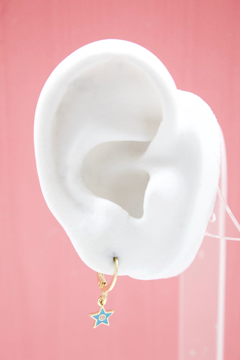 A white display ear showcases a gold huggie hoop earring featuring a charming blue star pendant against a soft pink background.