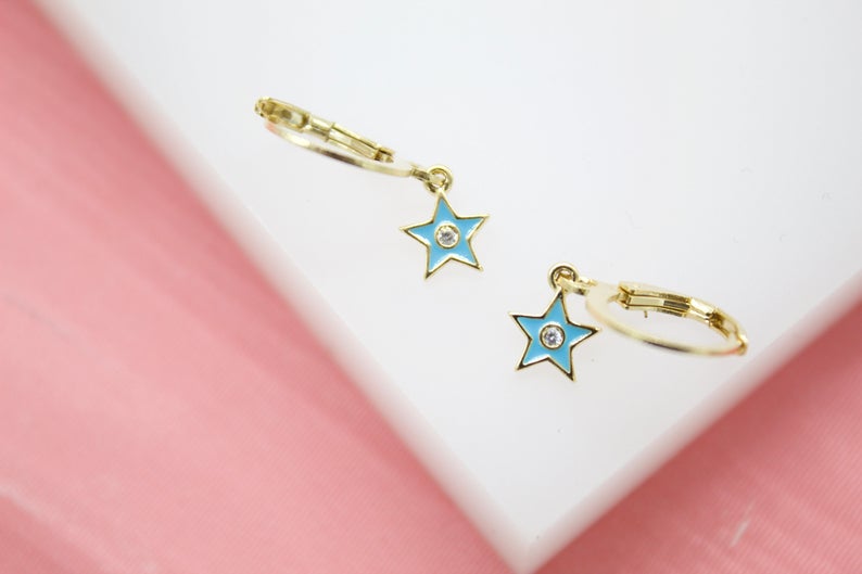 A pair of gold huggie hoop earrings features enamel blue star charms with center cubic zirconia stones resting on a white surface.