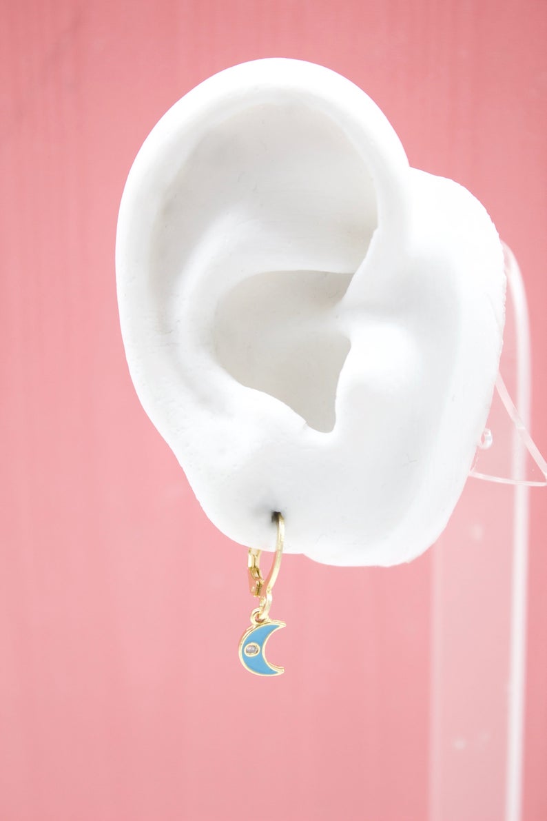 A gold hoop earring featuring a turquoise enamel crescent moon charm hangs on a white mannequin ear against a pink background.