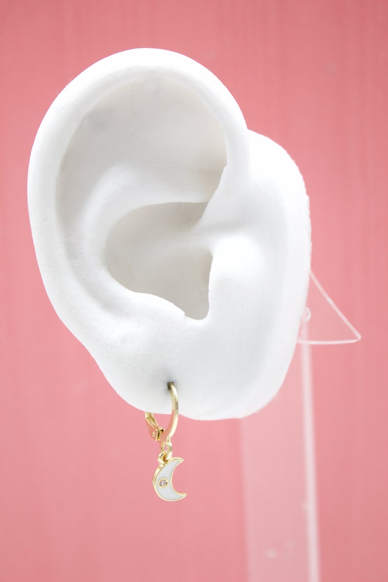 A white mannequin ear displays a gold hoop earring featuring a dangling white enamel crescent moon charm with a tiny gemstone.