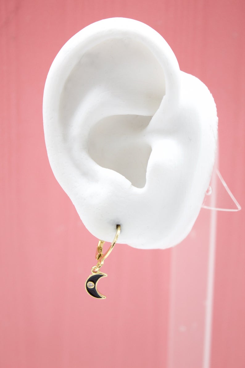 A white mannequin ear displays a gold huggie hoop earring featuring a black enamel crescent moon charm with a tiny diamond accent.