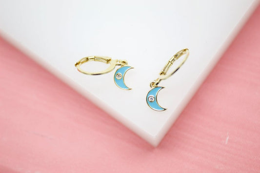 A pair of gold huggie hoop earrings featuring turquoise enamel crescent moon charms rests on a white display surface.